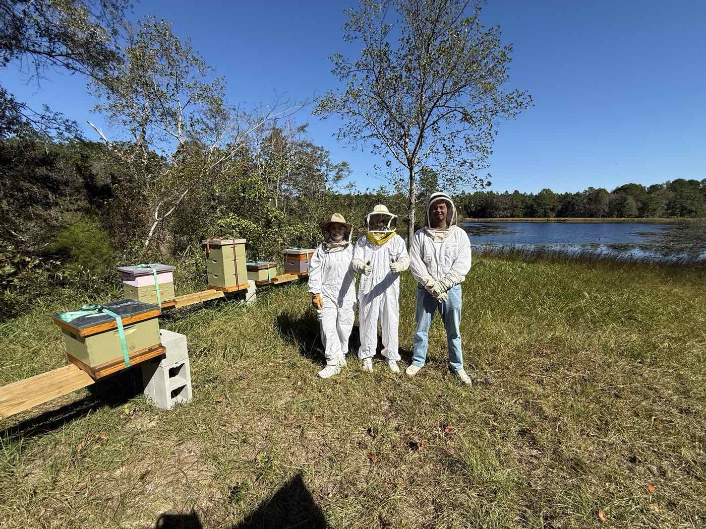 Three new beekeepers in bee suits giving a thumbs up beside the hives at a lakeside bee yard